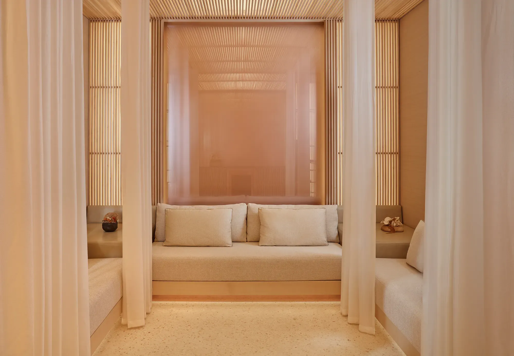 Serene wellness lounge with soft lighting, neutral tones, and cushioned benches framed by sheer curtains and wooden slats