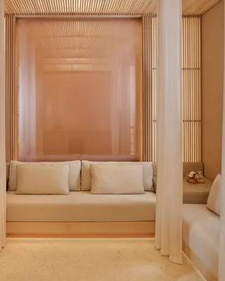 Serene wellness lounge with soft lighting, neutral tones, and cushioned benches framed by sheer curtains and wooden slats