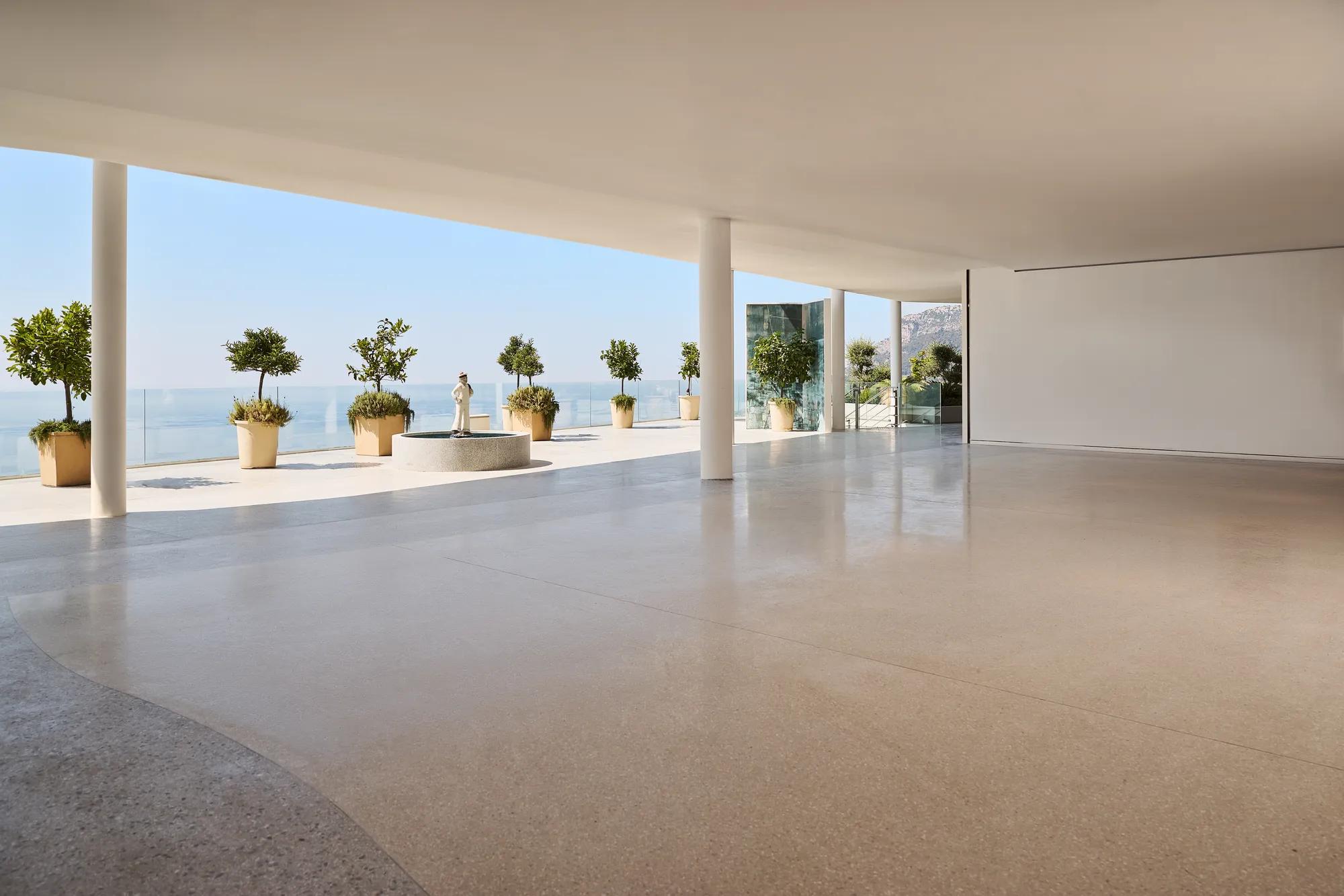 Modern open space with polished terrazzo flooring, white ceiling, and support columns, leading to a terrace overlooking the ocean. The terrace features large potted plants and a classical statue on a pedestal, with glass railings offering unobstructed sea views under a clear blue sky.
