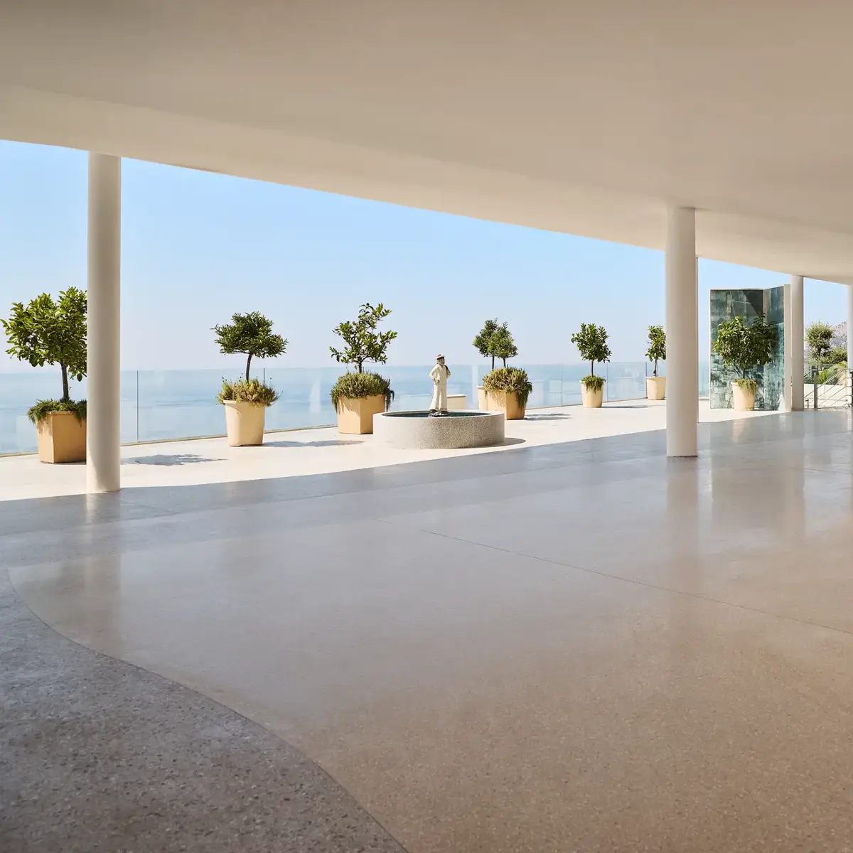 Modern open space with polished terrazzo flooring, white ceiling, and support columns, leading to a terrace overlooking the ocean. The terrace features large potted plants and a classical statue on a pedestal, with glass railings offering unobstructed sea views under a clear blue sky.