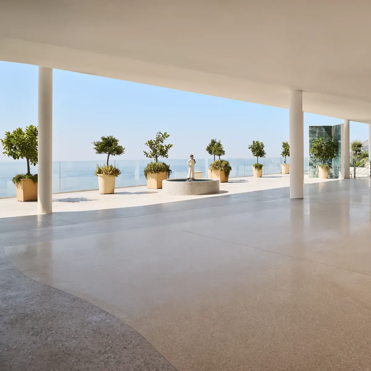 Modern open space with polished terrazzo flooring, white ceiling, and support columns, leading to a terrace overlooking the ocean. The terrace features large potted plants and a classical statue on a pedestal, with glass railings offering unobstructed sea views under a clear blue sky.