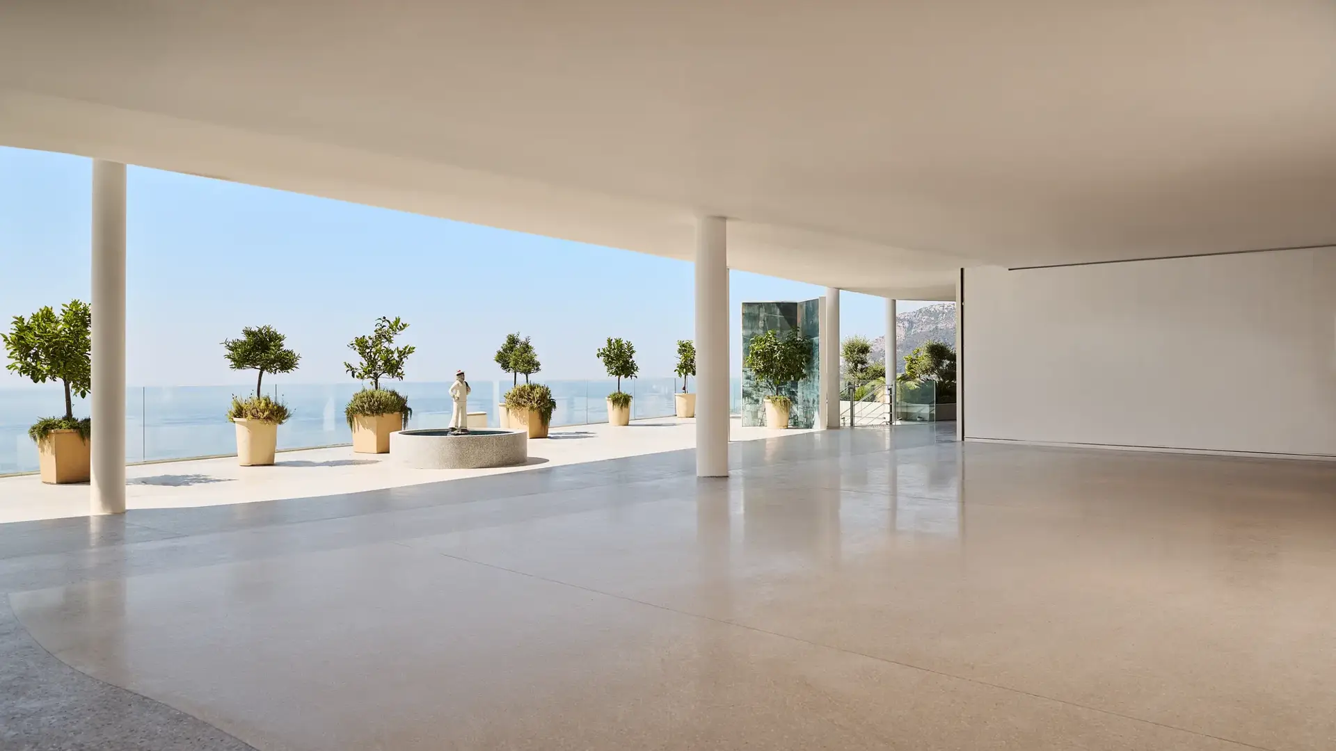 Modern open space with polished terrazzo flooring, white ceiling, and support columns, leading to a terrace overlooking the ocean. The terrace features large potted plants and a classical statue on a pedestal, with glass railings offering unobstructed sea views under a clear blue sky.