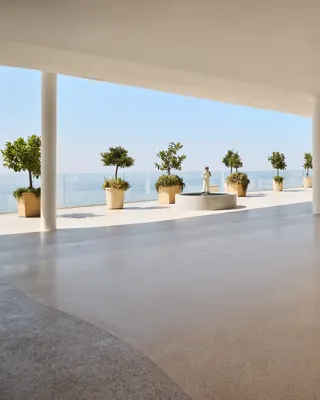Modern open space with polished terrazzo flooring, white ceiling, and support columns, leading to a terrace overlooking the ocean. The terrace features large potted plants and a classical statue on a pedestal, with glass railings offering unobstructed sea views under a clear blue sky.