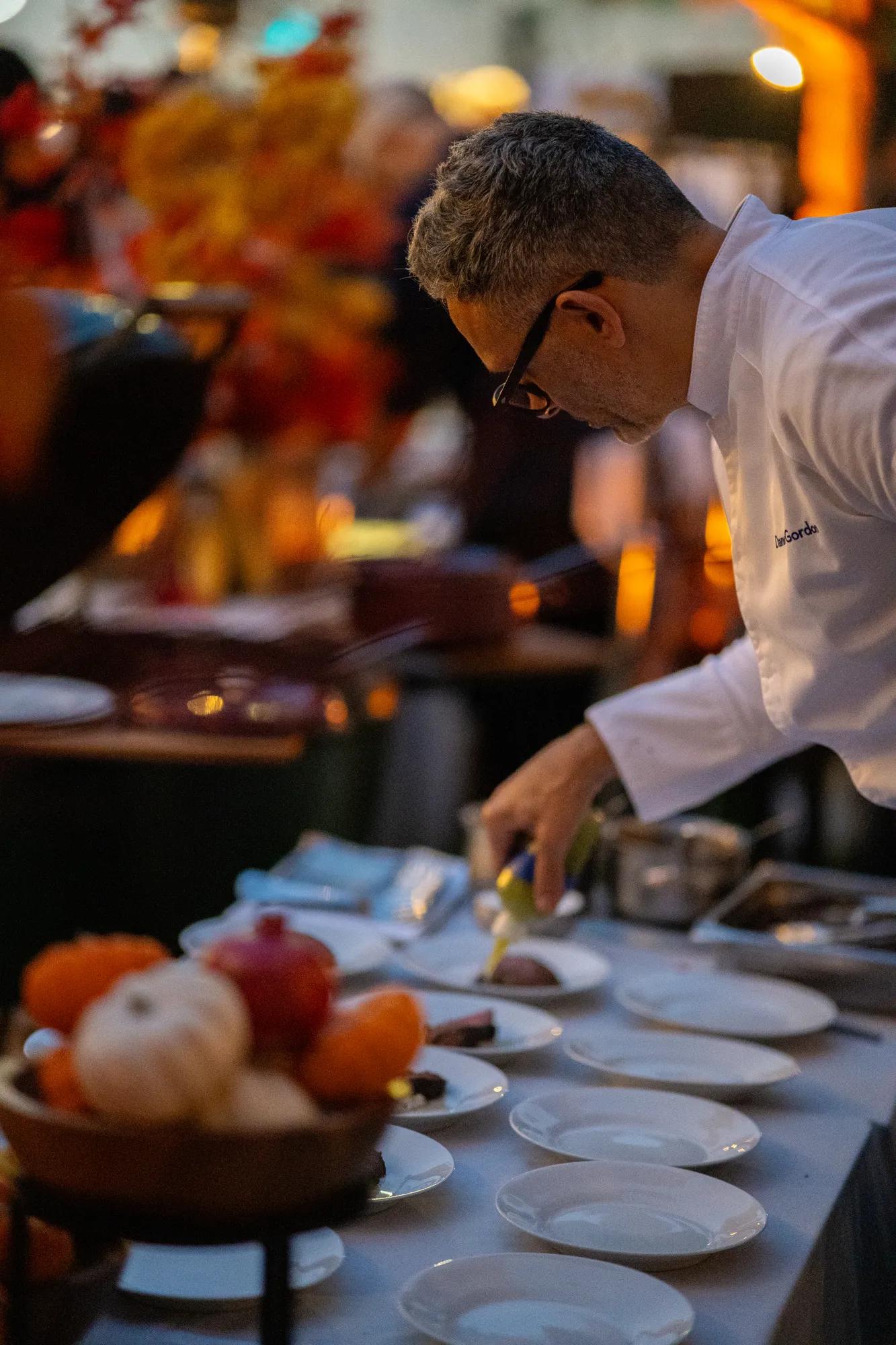 Chef plating and finishing dishes at a dining event with guests seated in the background.