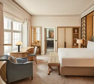Spacious bedroom with a king-size bed, geometric wood-panelled headboard, writing desk by a large window, and soft neutral furnishings in beige and blue tones.