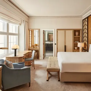 Spacious bedroom with a king-size bed, geometric wood-panelled headboard, writing desk by a large window, and soft neutral furnishings in beige and blue tones.