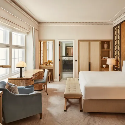 Spacious bedroom with a king-size bed, geometric wood-panelled headboard, writing desk by a large window, and soft neutral furnishings in beige and blue tones.