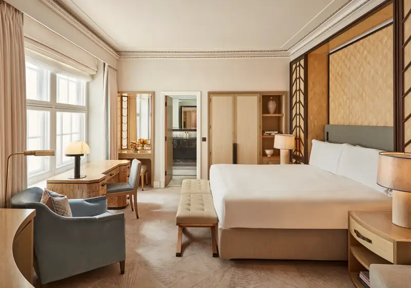 Spacious bedroom with a king-size bed, geometric wood-panelled headboard, writing desk by a large window, and soft neutral furnishings in beige and blue tones.