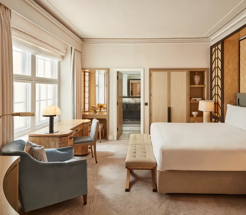 Spacious bedroom with a king-size bed, geometric wood-panelled headboard, writing desk by a large window, and soft neutral furnishings in beige and blue tones.