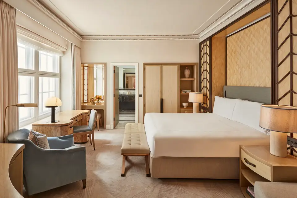 Spacious bedroom with a king-size bed, geometric wood-panelled headboard, writing desk by a large window, and soft neutral furnishings in beige and blue tones.