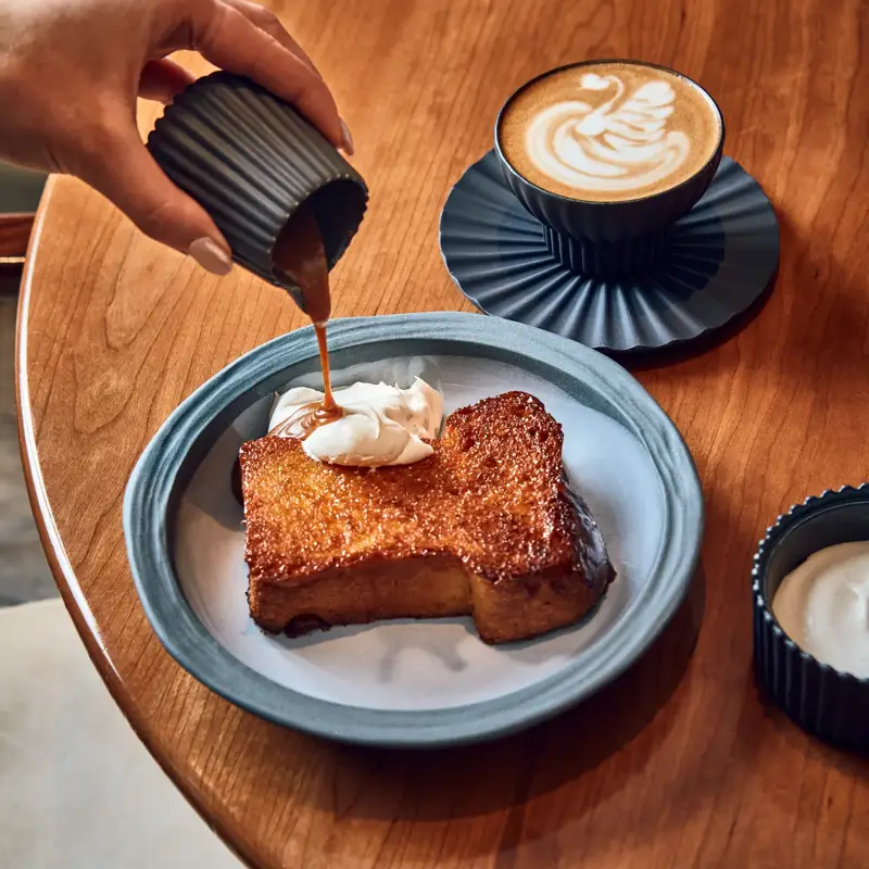 Hand pouring caramel sauce over brioche with cream, served with latte in blue cup.