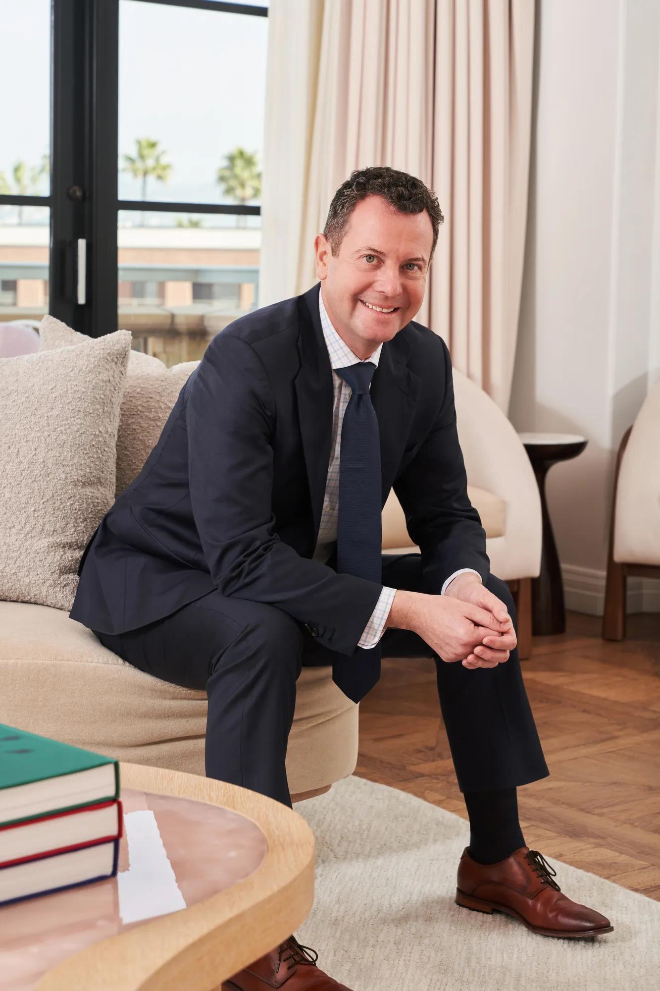 Person in a navy suit seated in a refined hotel lounge, with floor-to-ceiling windows, soft furnishings, and palm trees visible outside.