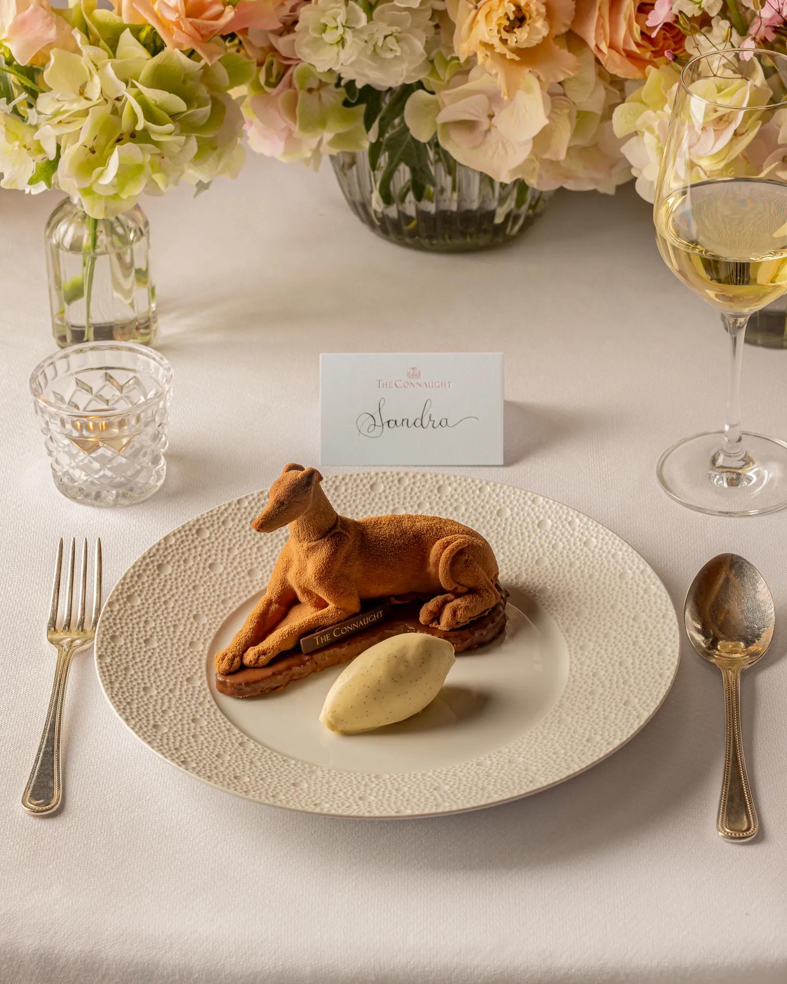 Plated dessert shaped like a reclining dog with a scoop of ice cream, set at an elegant place setting with flowers behind.