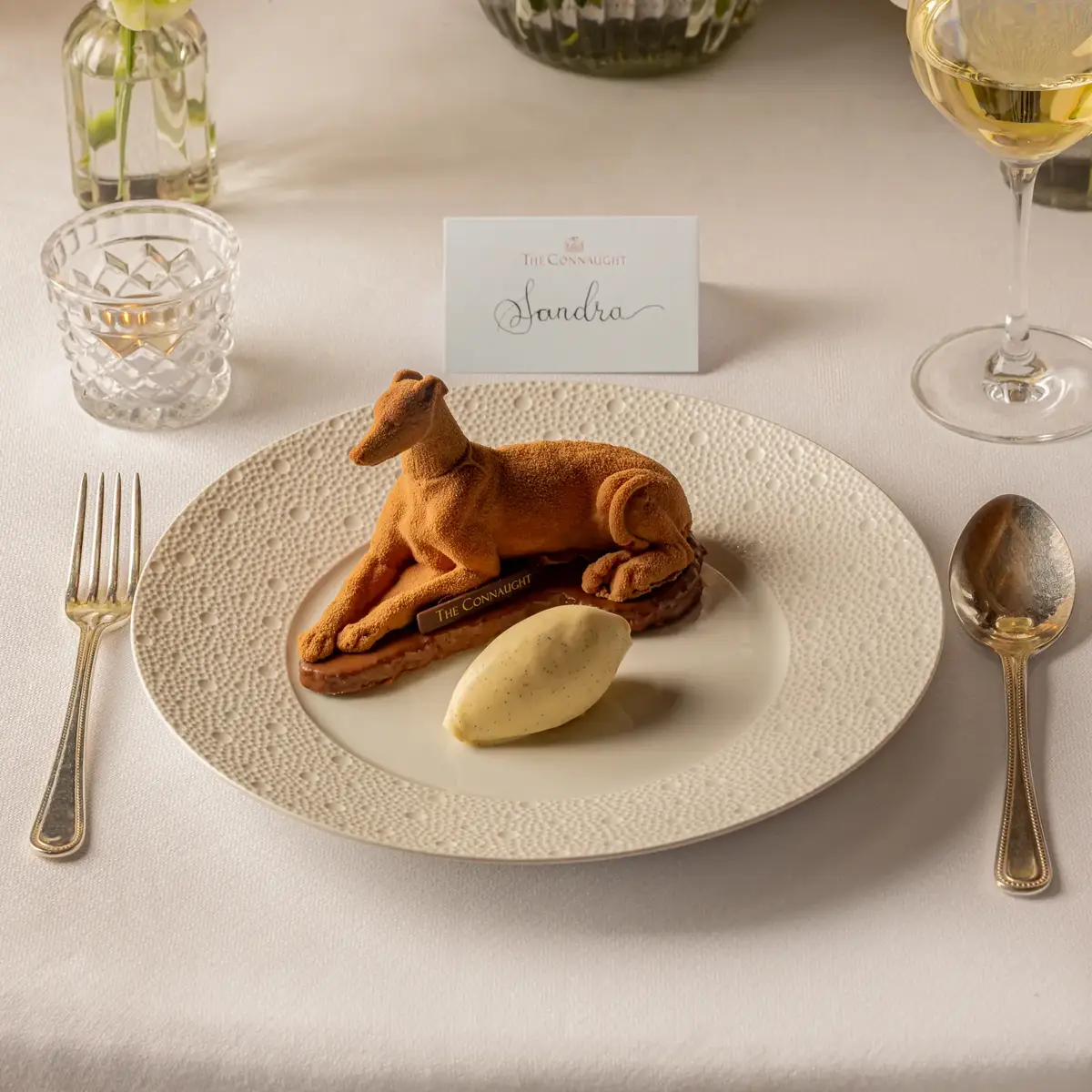 Plated dessert shaped like a reclining dog with a scoop of ice cream, set at an elegant place setting with flowers behind.