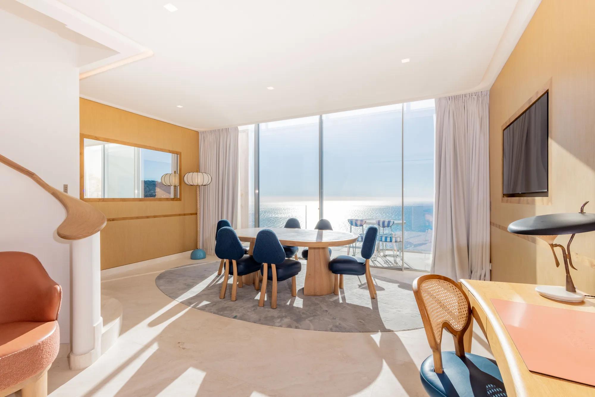 Salle à manger lumineuse avec table ronde en bois, chaises bleues et baies vitrées donnant sur la mer, agrémentée de finitions en bois chaleureux.