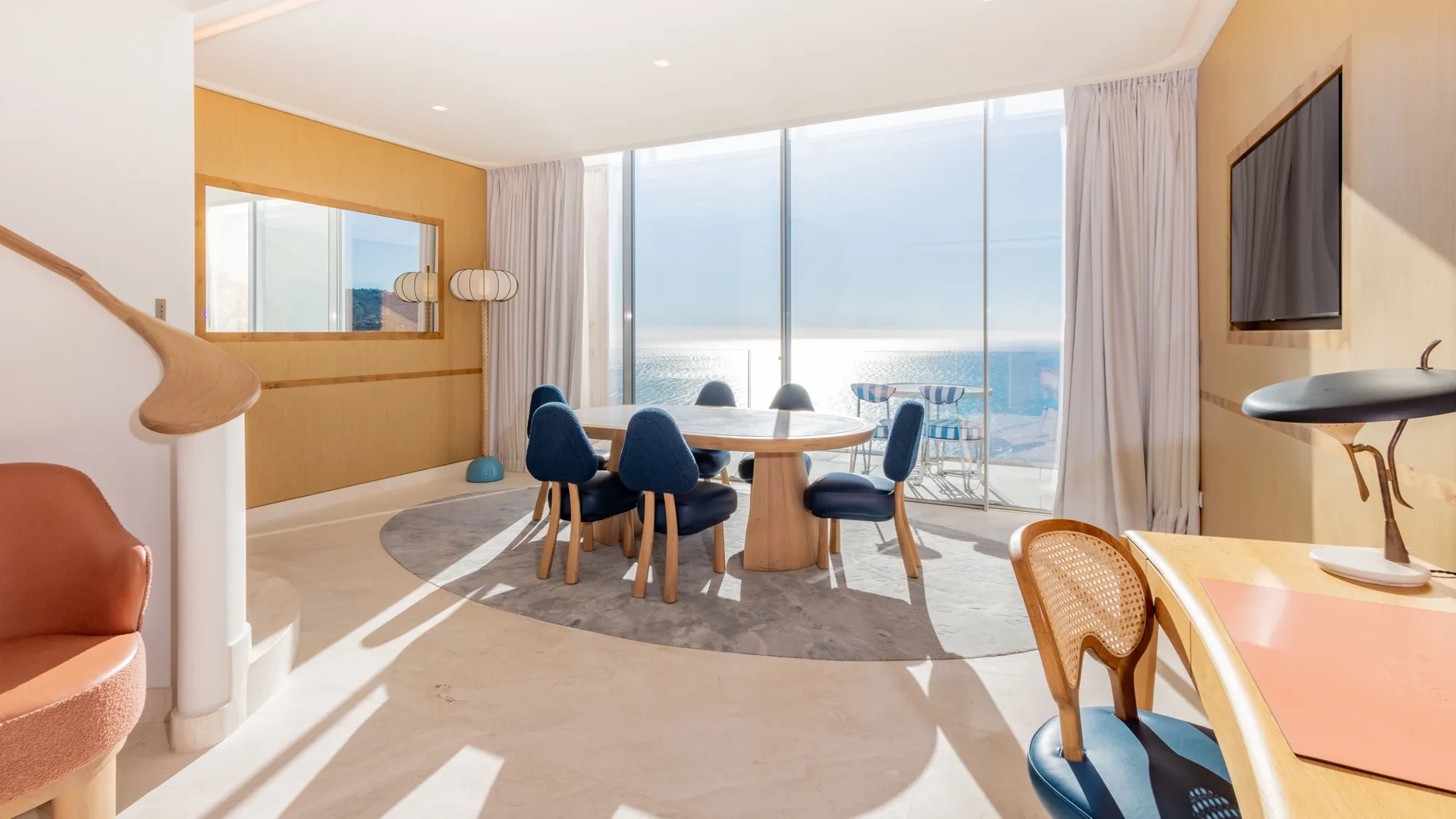 Salle à manger lumineuse avec table ronde en bois, chaises bleues et baies vitrées donnant sur la mer, agrémentée de finitions en bois chaleureux.