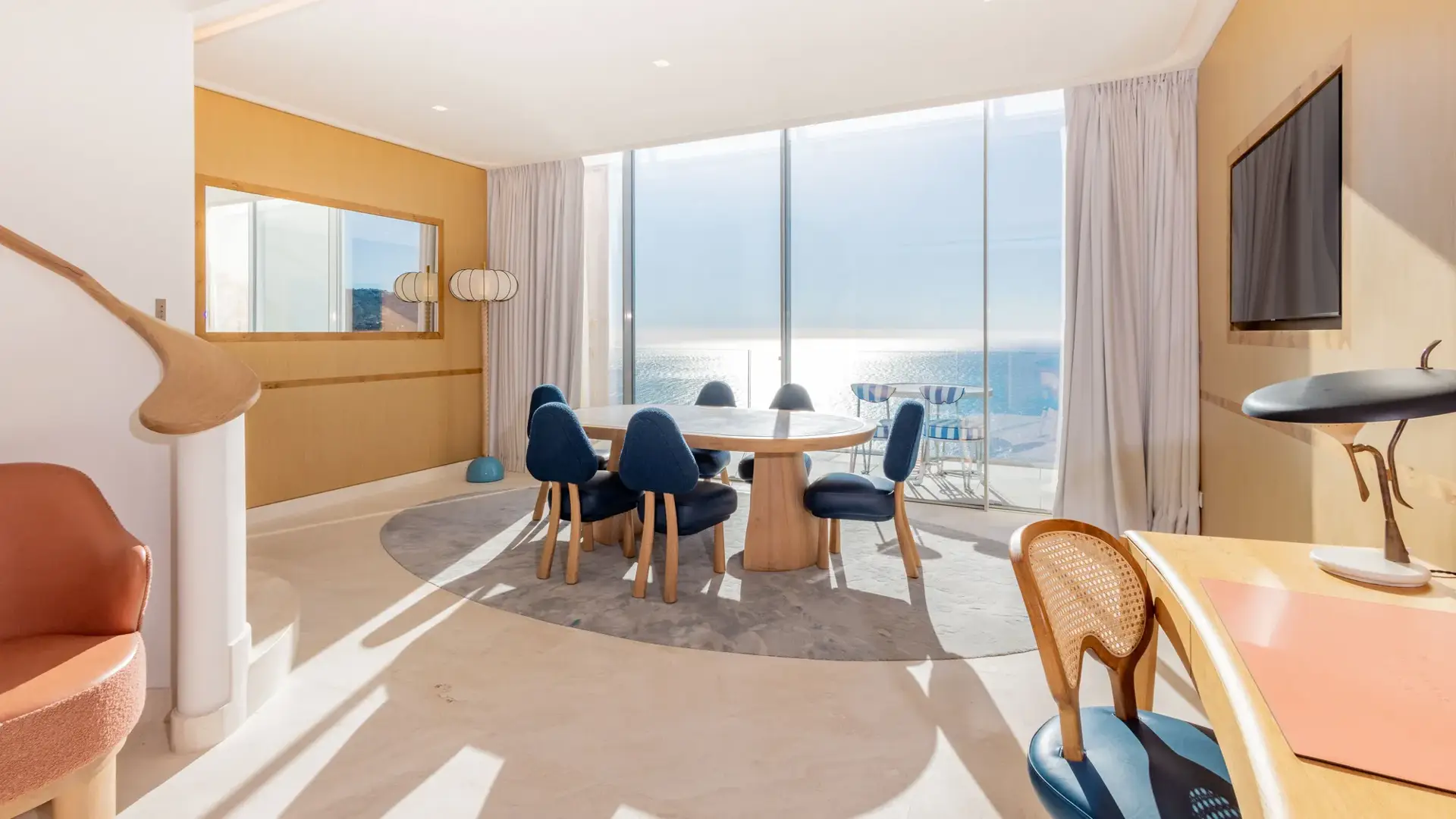 Salle à manger lumineuse avec table ronde en bois, chaises bleues et baies vitrées donnant sur la mer, agrémentée de finitions en bois chaleureux.