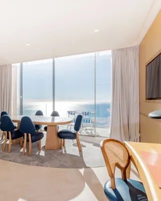 Salle à manger lumineuse avec table ronde en bois, chaises bleues et baies vitrées donnant sur la mer, agrémentée de finitions en bois chaleureux.