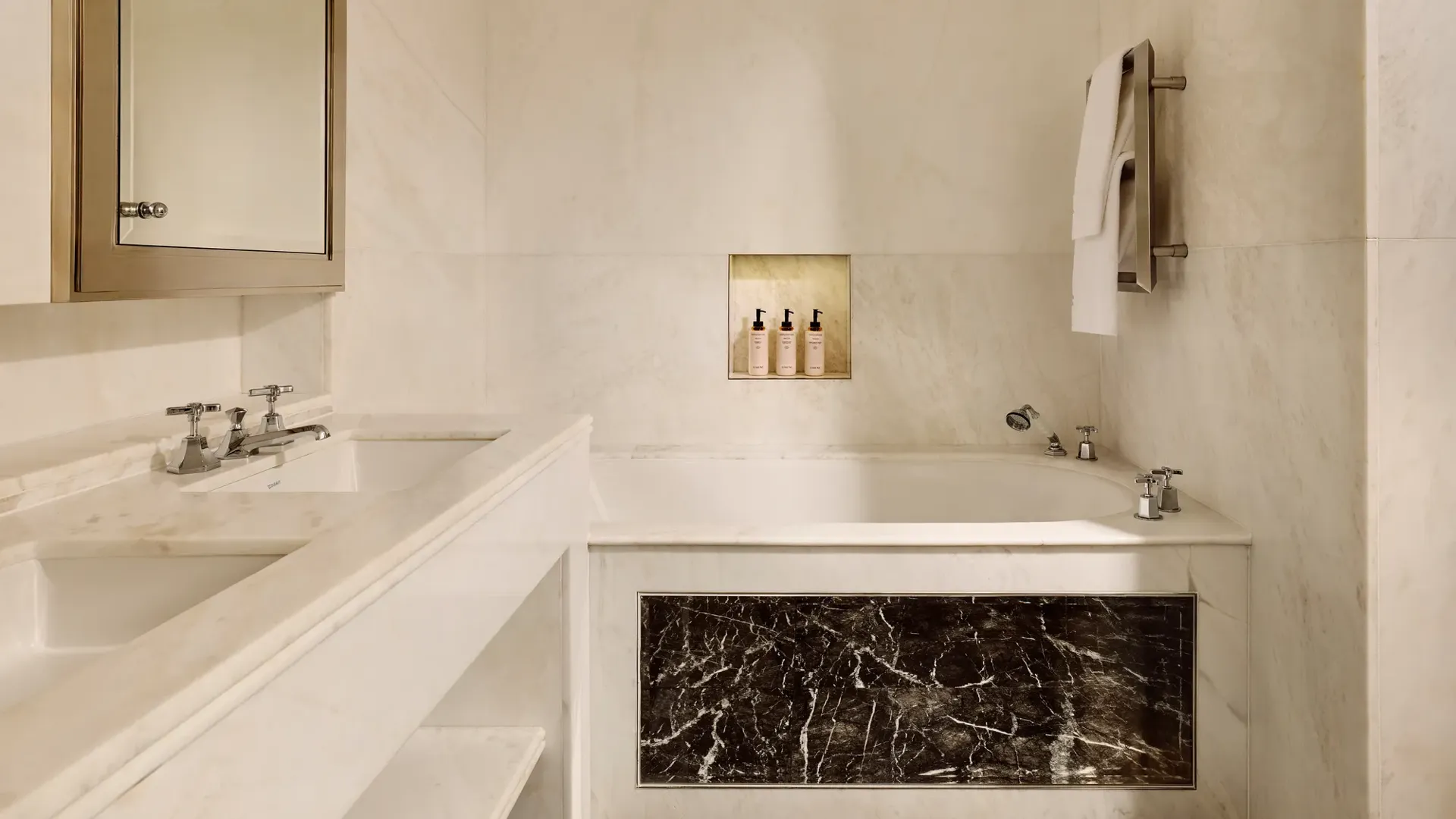 Luxurious marble bathroom with built-in bathtub, dark marble panel, double sink, and niche holding toiletries.