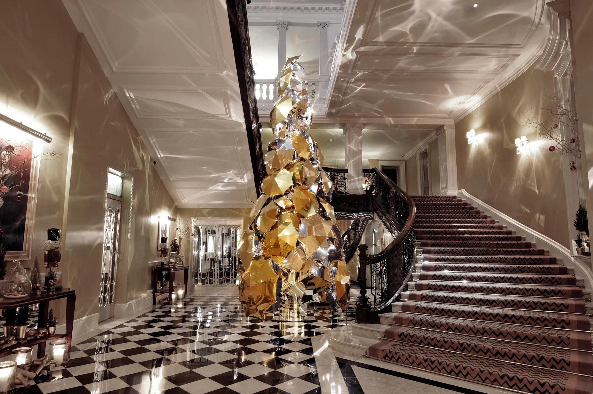 Grand hotel lobby with sweeping staircase and checkered floor, featuring a tall sculptural gold tree reflecting light across walls and ceiling.