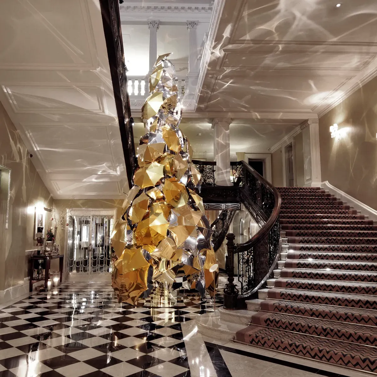 Grand hotel lobby with sweeping staircase and checkered floor, featuring a tall sculptural gold tree reflecting light across walls and ceiling.