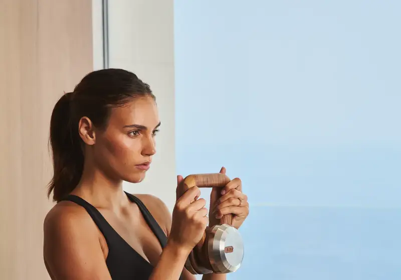 Woman in black sportswear holding a dumbbell upright near her chest, standing indoors by a window overlooking the sea.