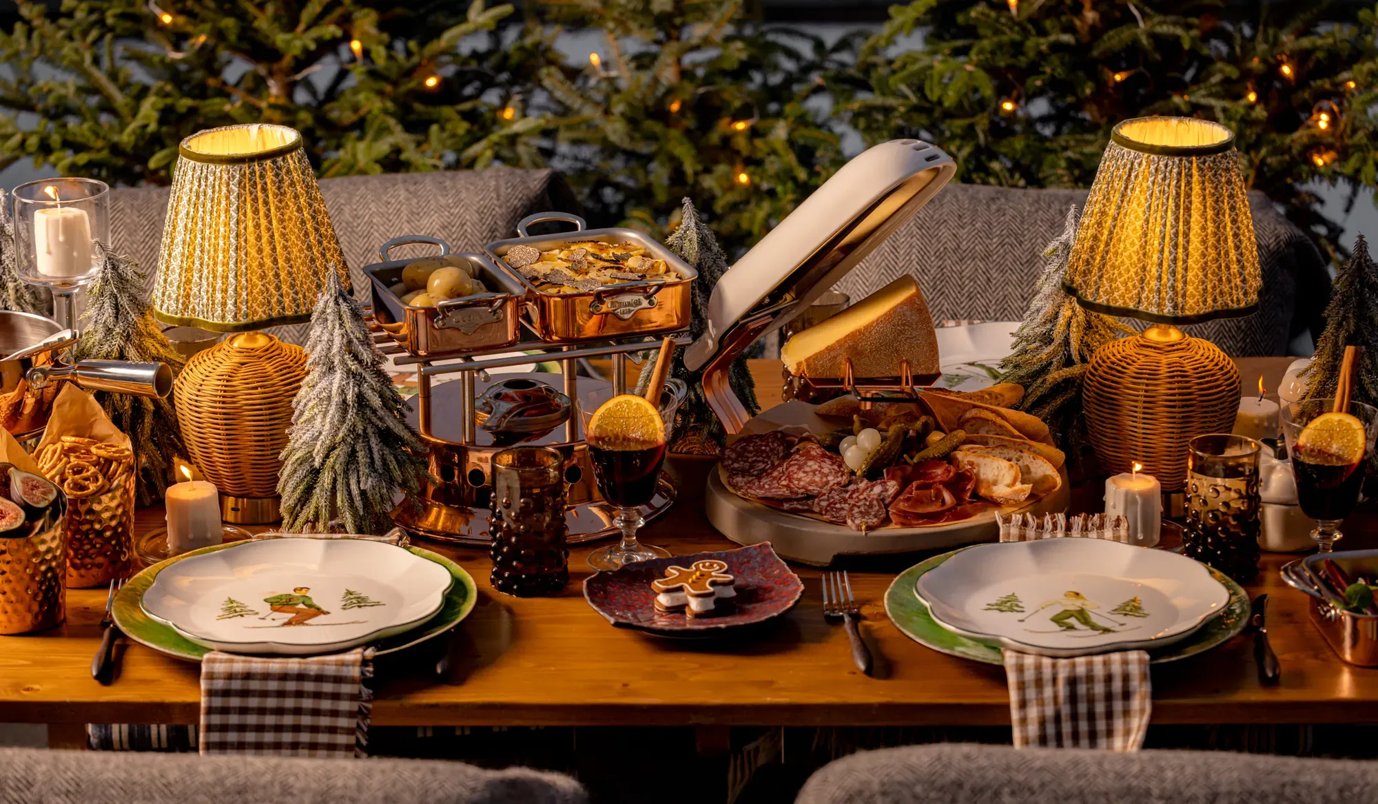 Festive dining table set with fondue, cured meats, candles, and small decorated trees, creating a warm holiday atmosphere