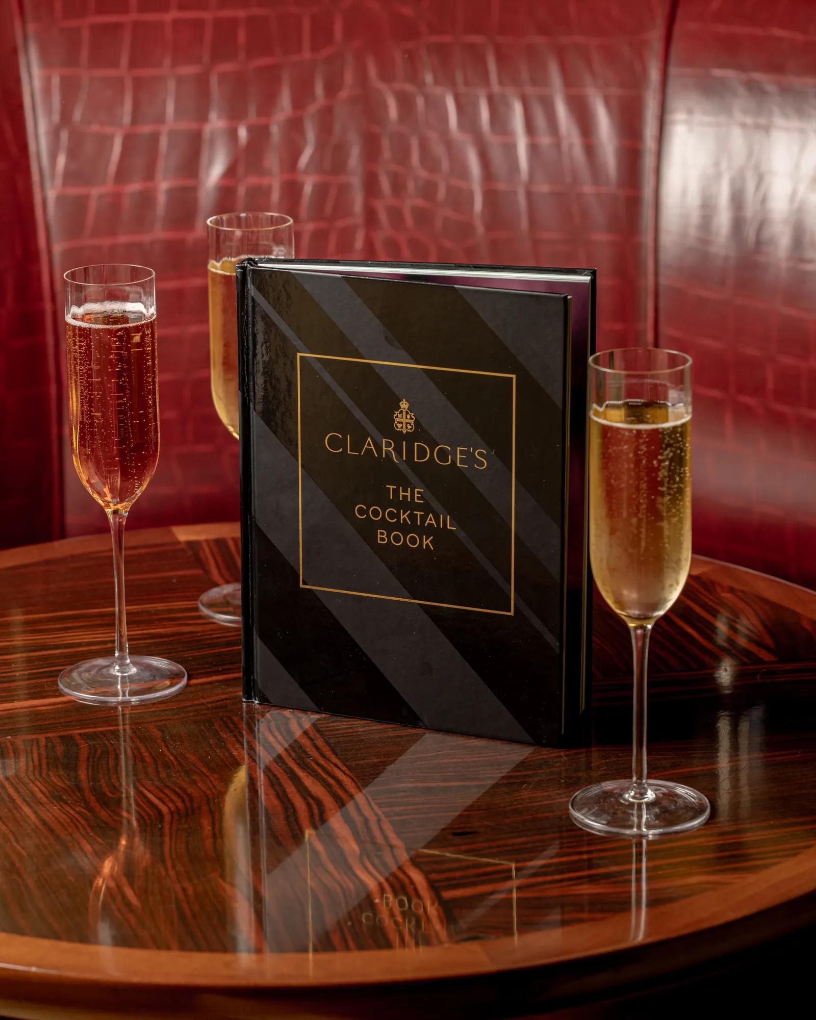 Claridge’s cocktail book on polished wooden table with three champagne flutes against red leather banquette