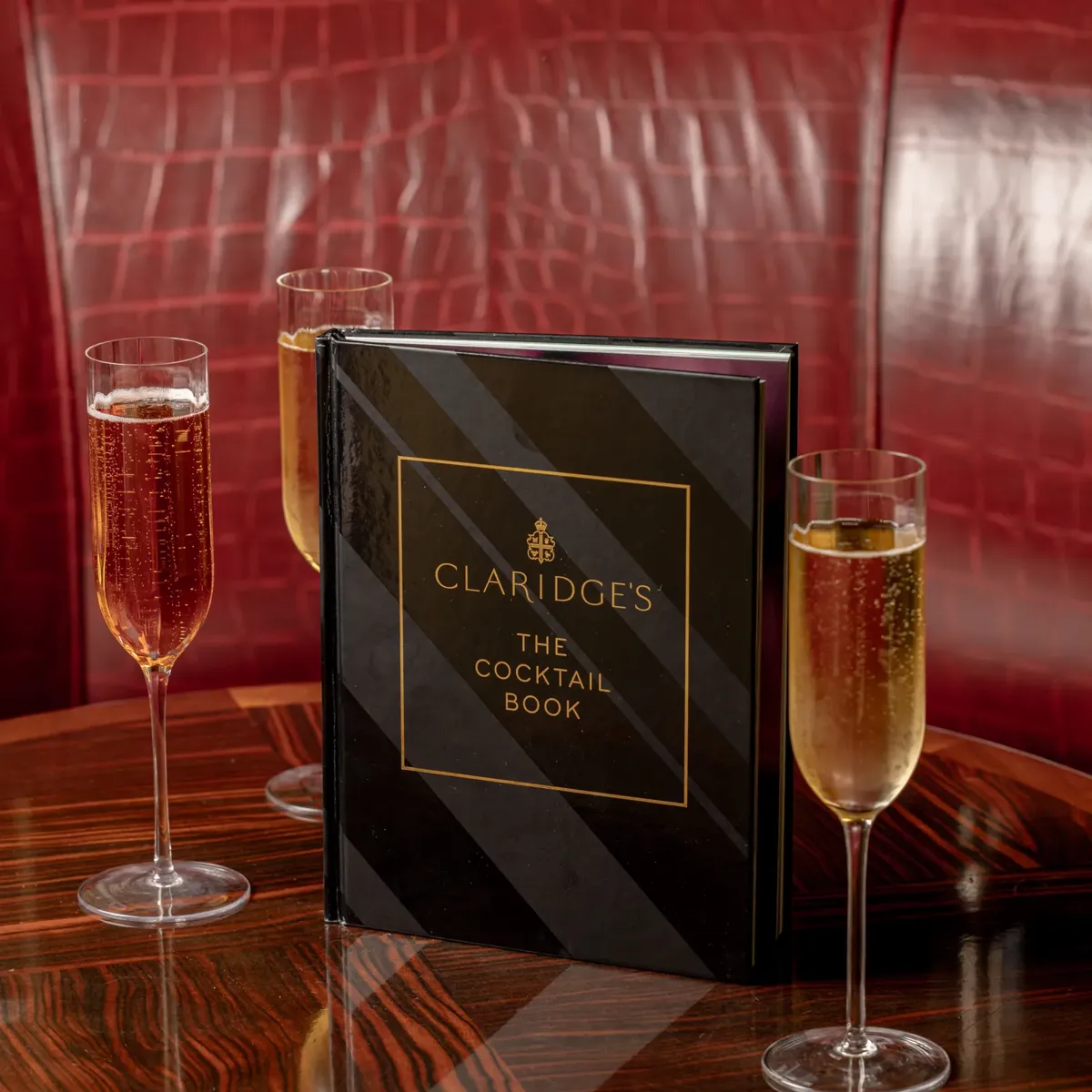 Claridge’s cocktail book on polished wooden table with three champagne flutes against red leather banquette