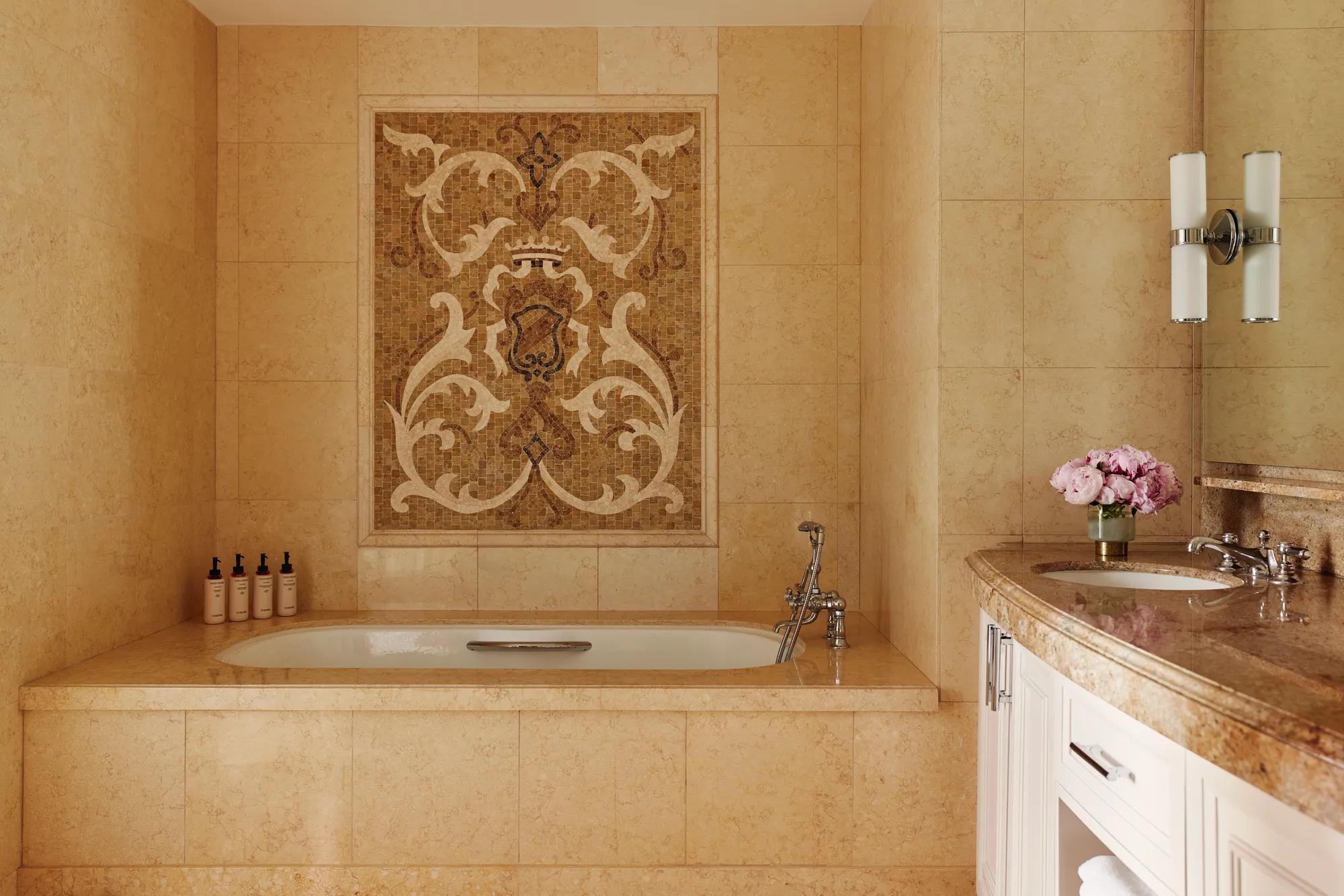Stone‑tiled bathroom with deep tub beneath an ornate mosaic panel and curved double‑sink vanity.