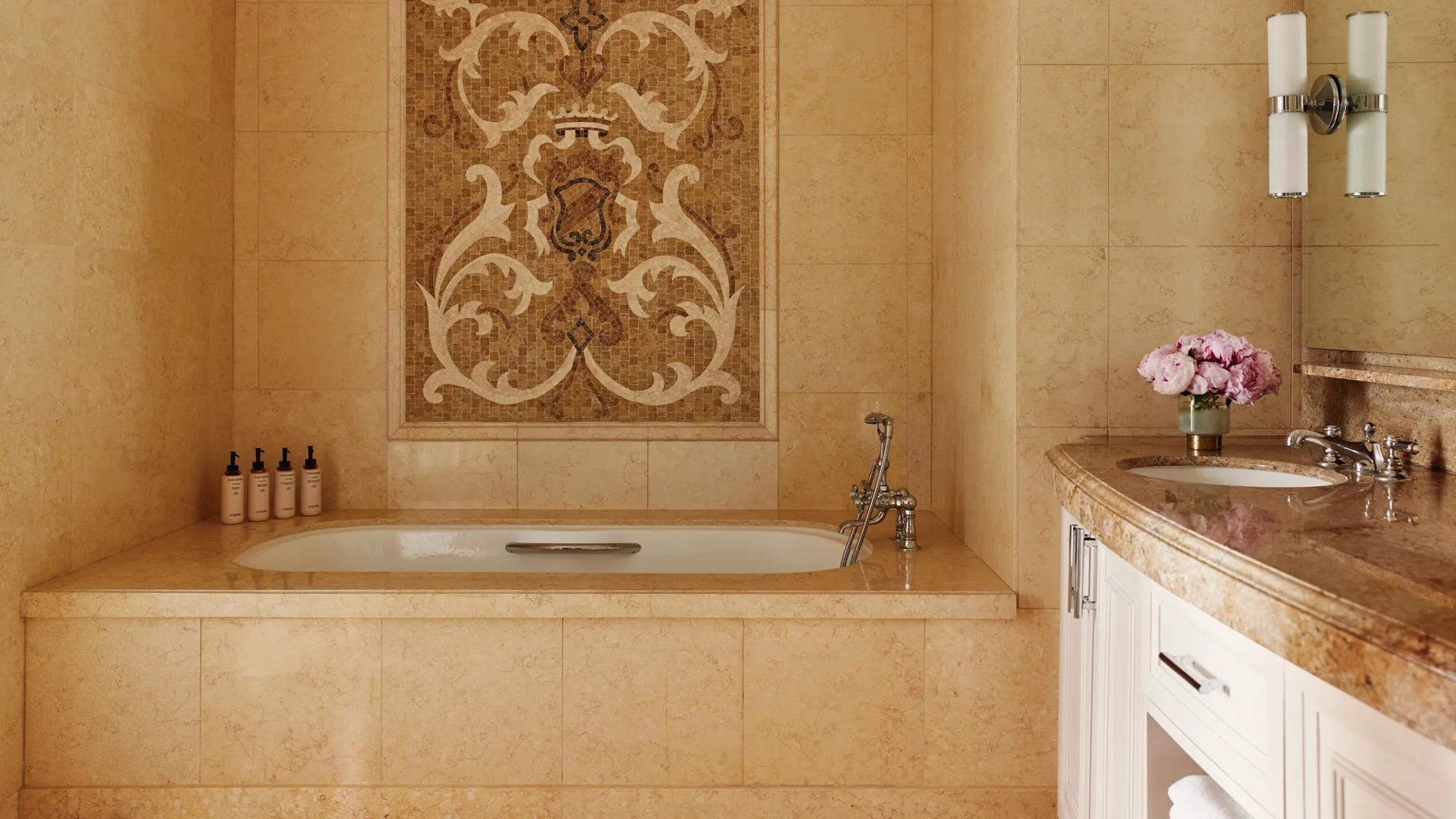Stone‑tiled bathroom with deep tub beneath an ornate mosaic panel and curved double‑sink vanity.