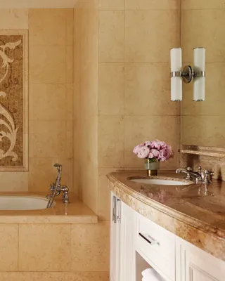 Stone‑tiled bathroom with deep tub beneath an ornate mosaic panel and curved double‑sink vanity.