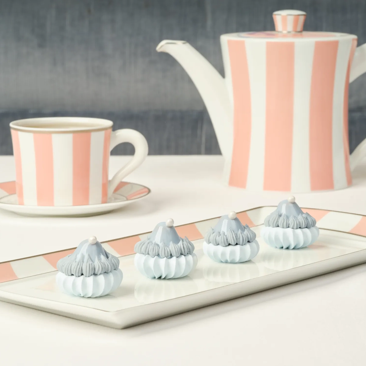 Striped pink-and-white teapot set with four blue-and-white meringue treats on a tray.