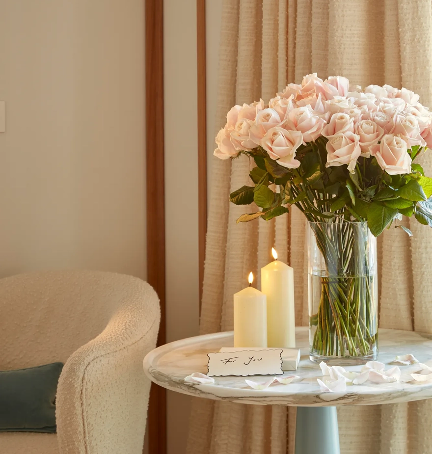 Blush pink roses in a glass vase beside two lit candles and a handwritten note on a small round table by the window.