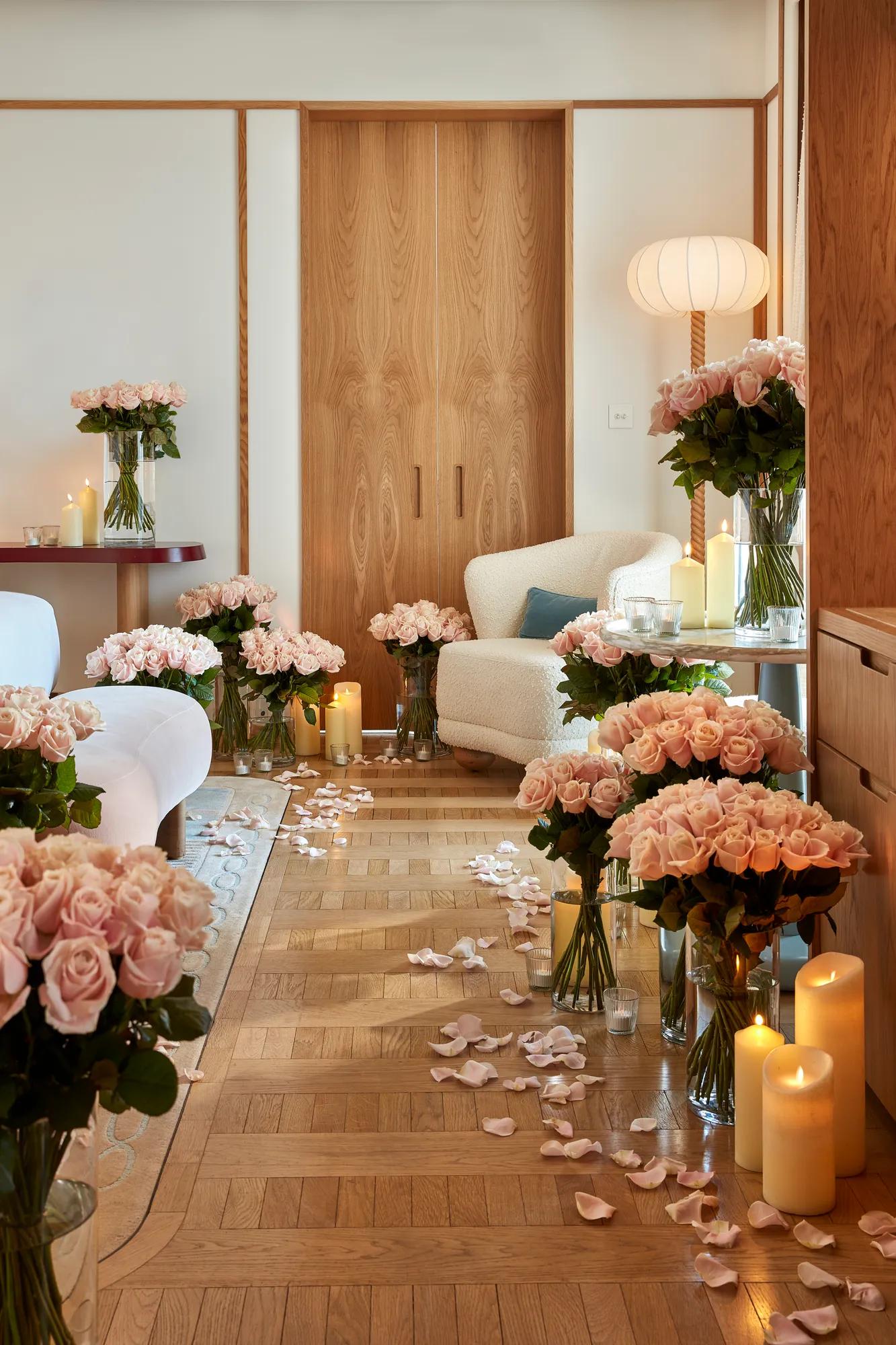 A romantic suite decorated with numerous vases of pale pink roses, glowing candles, and scattered rose petals on a parquet floor, with a cream armchair and soft lighting creating a warm, intimate atmosphere.