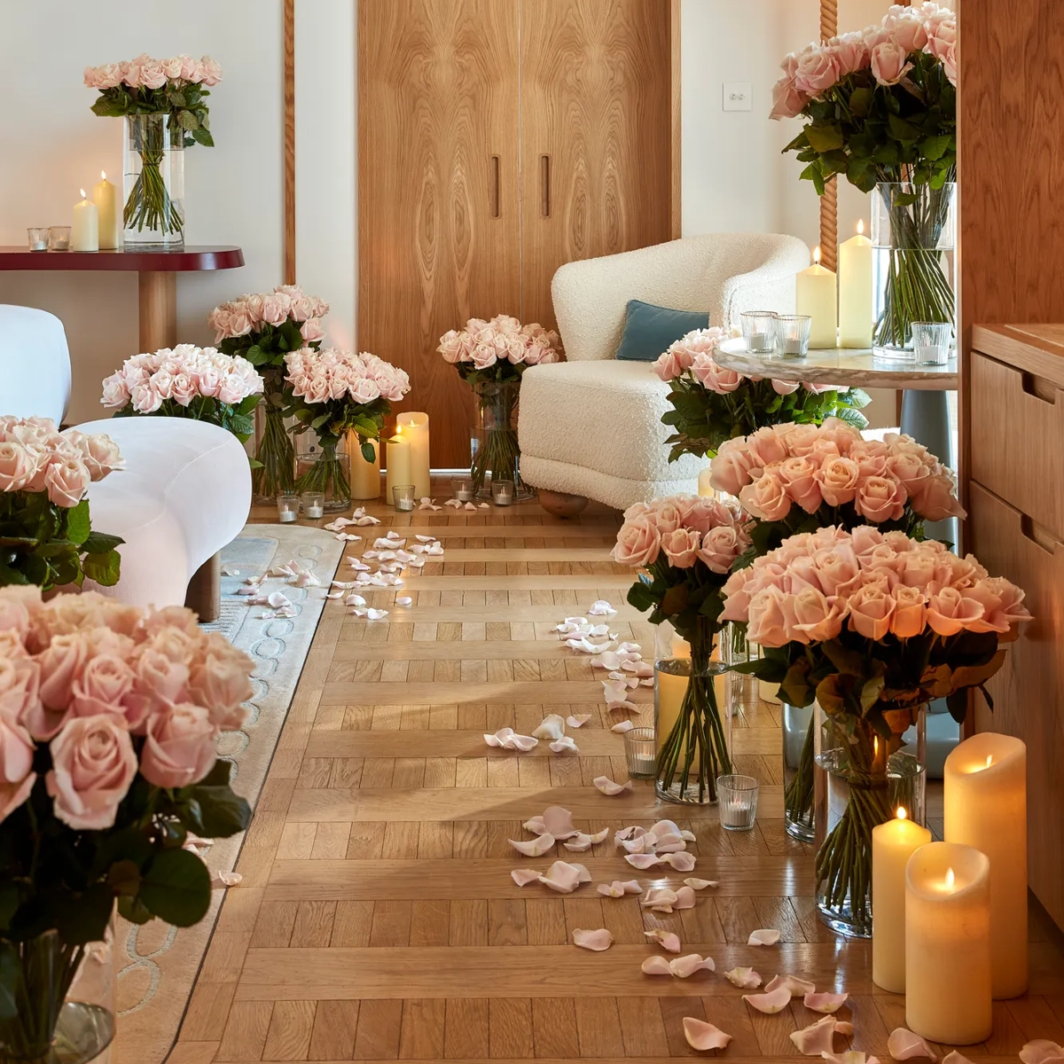 A romantic suite decorated with numerous vases of pale pink roses, glowing candles, and scattered rose petals on a parquet floor, with a cream armchair and soft lighting creating a warm, intimate atmosphere.