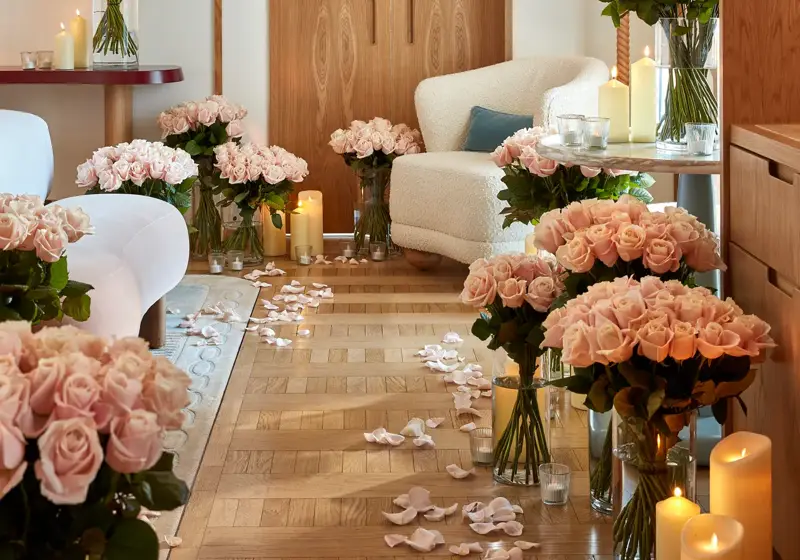 A romantic suite decorated with numerous vases of pale pink roses, glowing candles, and scattered rose petals on a parquet floor, with a cream armchair and soft lighting creating a warm, intimate atmosphere.