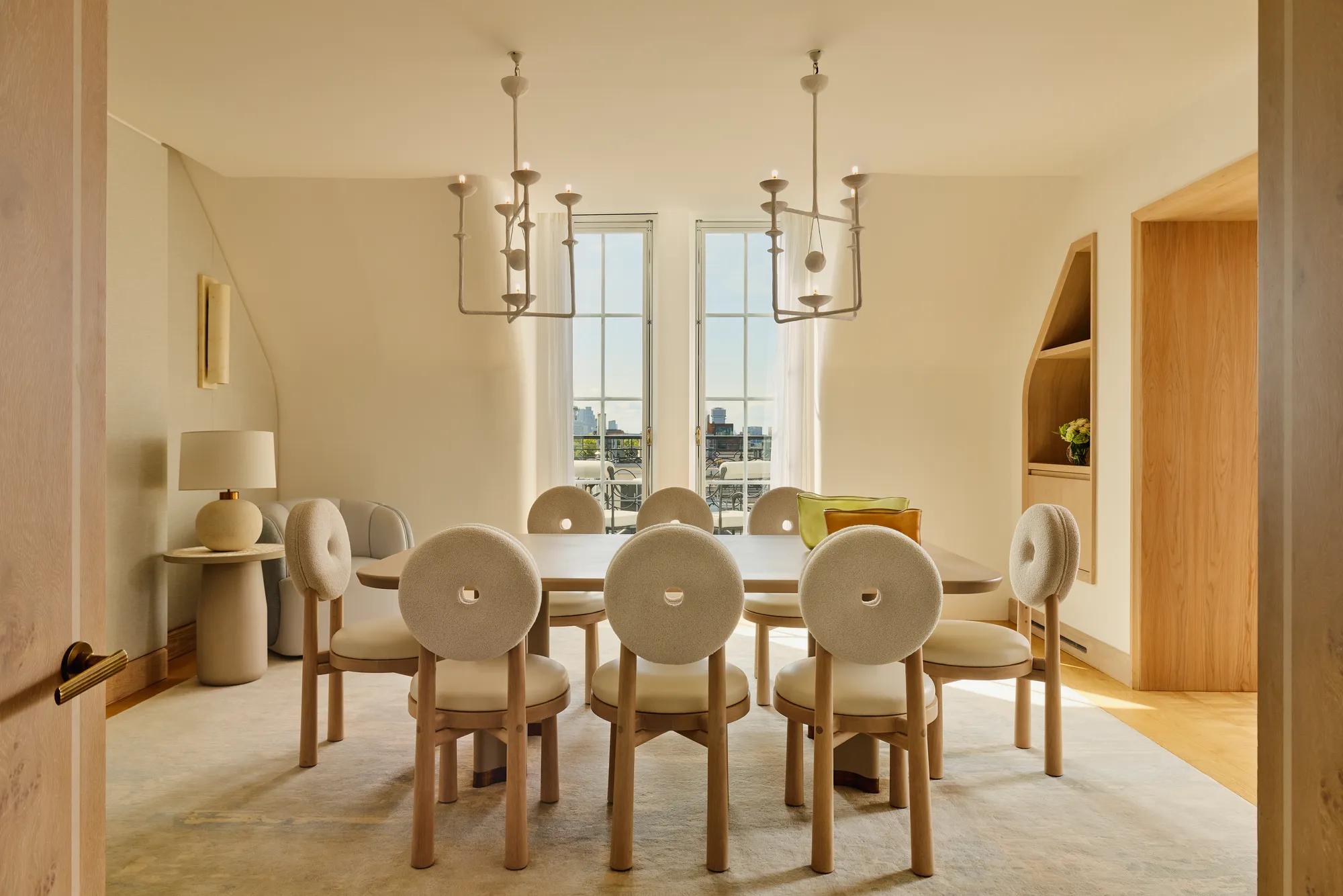 Light-filled dining room with a wooden table surrounded by eight round-backed cream chairs, featuring sculptural pendant lights and French doors opening to city views.