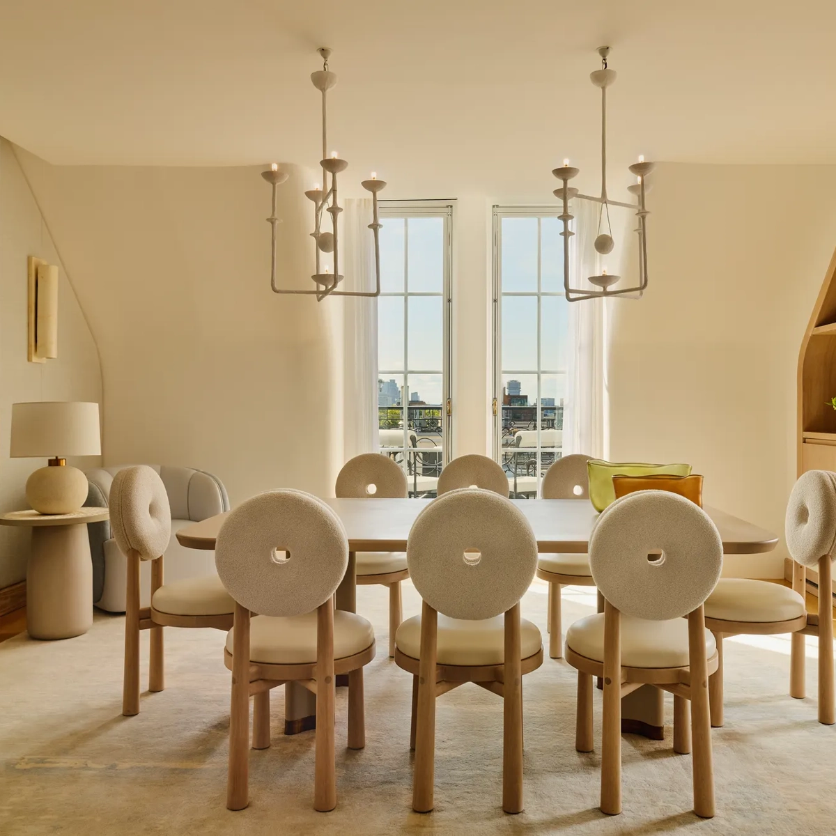 Light-filled dining room with a wooden table surrounded by eight round-backed cream chairs, featuring sculptural pendant lights and French doors opening to city views.