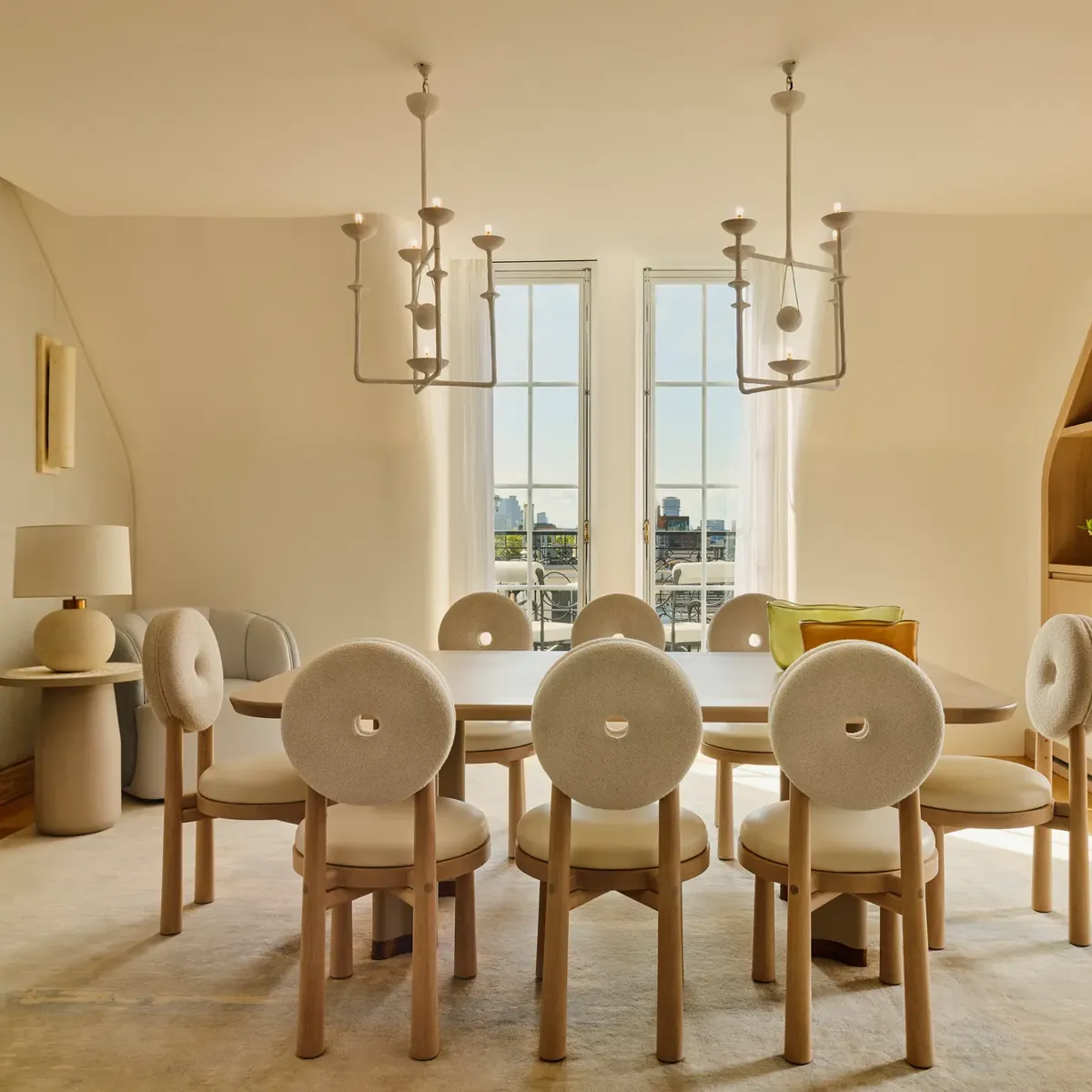 Light-filled dining room with a wooden table surrounded by eight round-backed cream chairs, featuring sculptural pendant lights and French doors opening to city views.