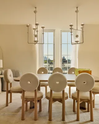 Light-filled dining room with a wooden table surrounded by eight round-backed cream chairs, featuring sculptural pendant lights and French doors opening to city views.