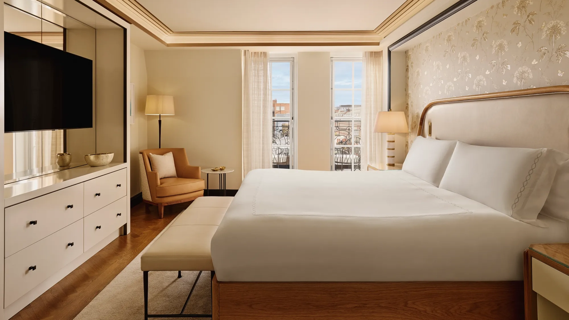 Serene bedroom featuring a king bed with a cream upholstered headboard, soft gold patterned wallpaper, a seating area with an armchair, and floor-to-ceiling windows letting in natural light.