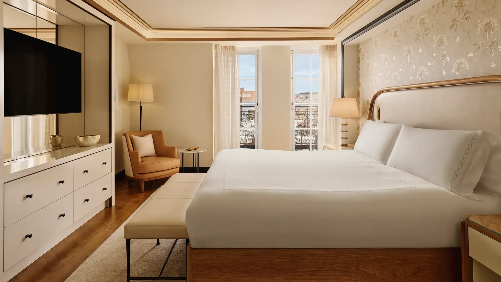 Serene bedroom featuring a king bed with a cream upholstered headboard, soft gold patterned wallpaper, a seating area with an armchair, and floor-to-ceiling windows letting in natural light.