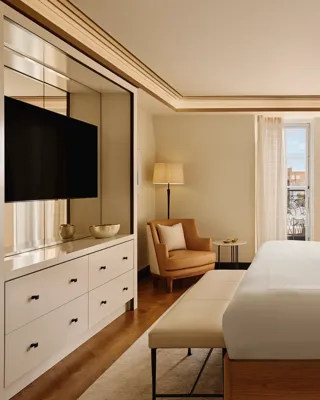 Serene bedroom featuring a king bed with a cream upholstered headboard, soft gold patterned wallpaper, a seating area with an armchair, and floor-to-ceiling windows letting in natural light.