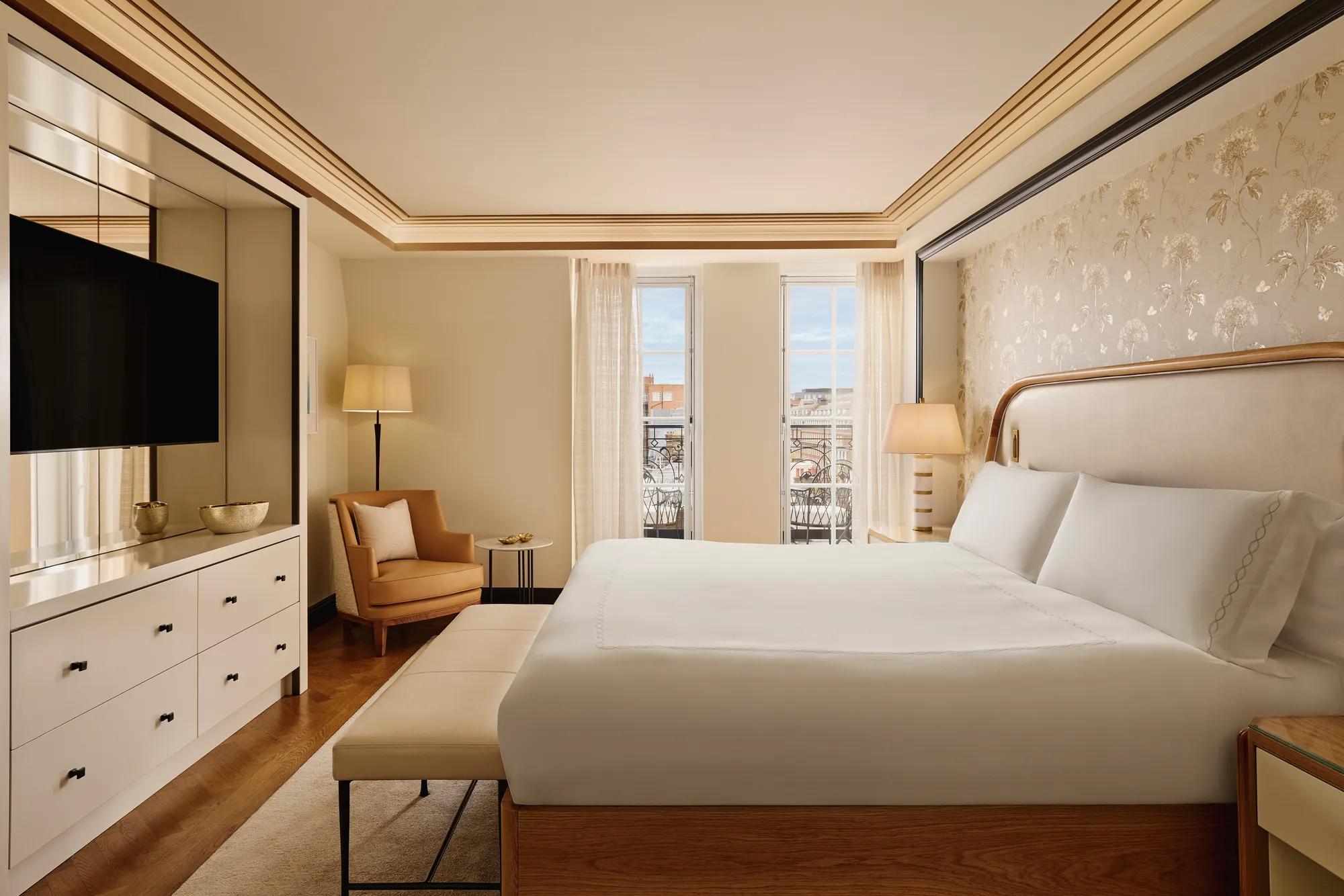 Serene bedroom featuring a king bed with a cream upholstered headboard, soft gold patterned wallpaper, a seating area with an armchair, and floor-to-ceiling windows letting in natural light.