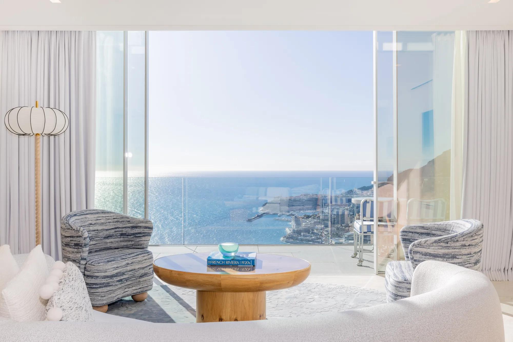 Salon lumineux avec canapé incurvé, fauteuils à motifs et table ronde en bois, s’ouvrant sur une terrasse avec vue panoramique sur la mer et la ville.