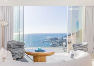 Salon lumineux avec canapé incurvé, fauteuils à motifs et table ronde en bois, s’ouvrant sur une terrasse avec vue panoramique sur la mer et la ville.