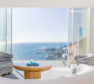 Salon lumineux avec canapé incurvé, fauteuils à motifs et table ronde en bois, s’ouvrant sur une terrasse avec vue panoramique sur la mer et la ville.