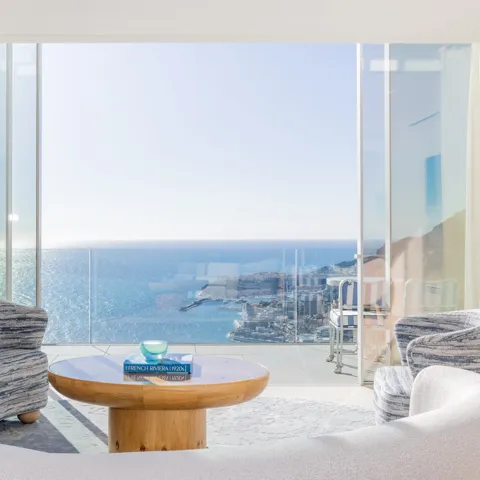 Bright living room with curved sofa, patterned chairs, and round wooden table, opening onto a terrace with panoramic sea and city views.