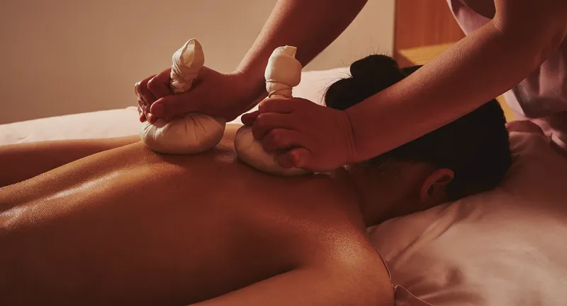 Spa therapist applying heated herbal compresses to a guest’s back during a massage treatment.